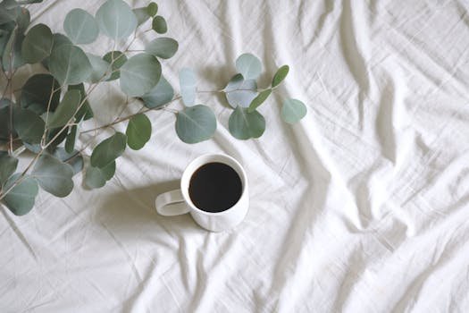 A simple arrangement of coffee with eucalyptus leaves on a white bedsheet.